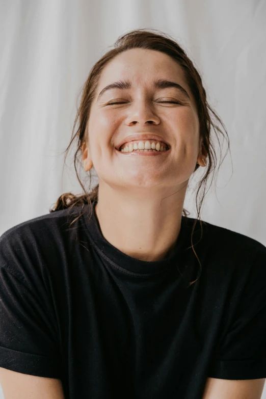 a woman is smiling with her eyes closed, by irakli nadar, happening, plain background, wearing a black t-shirt, head bent back in laughter, goofy smile