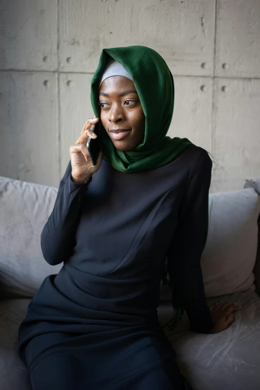 a woman sitting on a couch talking on a cell phone, inspired by Maryam Hashemi, trending on pexels, hurufiyya, green dress with a black hood, black female, black scarf, professional modeling