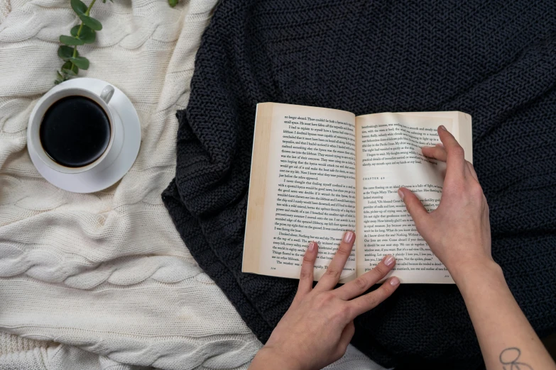 a woman reading a book next to a cup of coffee, pexels contest winner, private press, black wool sweater, wearing nanotech honeycomb robe, lined in cotton, hands reaching for her