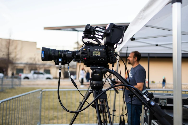 a man standing next to a camera on a tripod, sports broadcast, canon pro, cooke anamorphic / i lenses, live 3hr festival videoclip