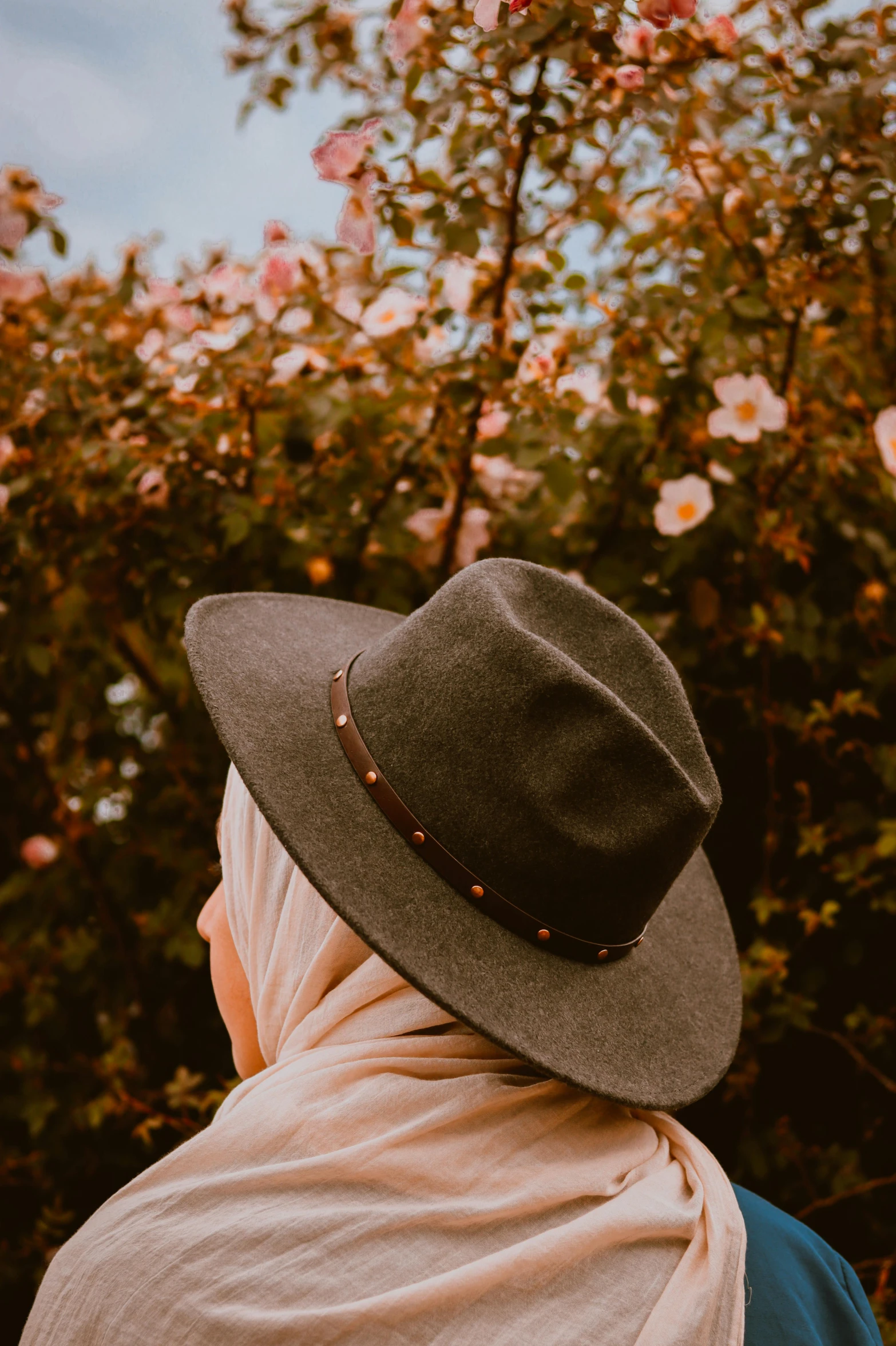 a woman wearing a hat and a scarf, trending on pexels, flowers in background, desaturated, back - shot, fedora