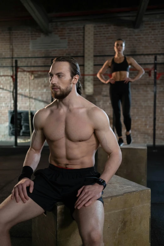 a man sitting on top of a box next to a woman, sweaty abs, background a gym, promo image, male hero