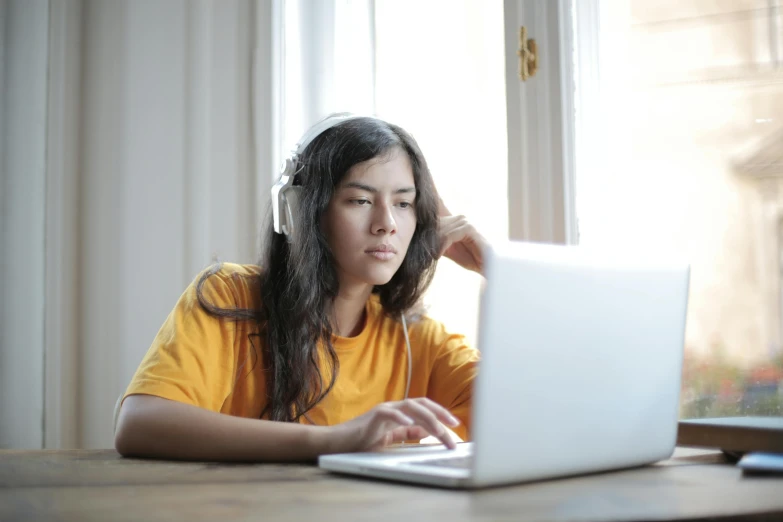 a woman sitting at a table using a laptop computer, trending on pexels, wearing a yellow hoodie, with head phones, pokimane, student