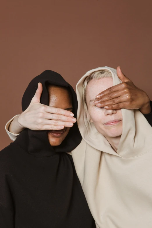 two women covering their eyes with their hands, by Matija Jama, hurufiyya, beige hoodie, wearing a black cloak, enes dirig, diversity