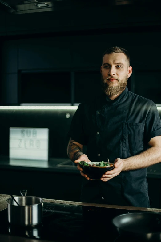 a man standing in a kitchen holding a bowl of food, with a beard and a black shirt, style of seb mckinnon, plating, commercially ready