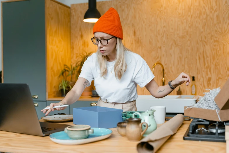 a woman in an orange hat working on a laptop, by Julia Pishtar, trending on pexels, knick knacks, delivering parsel box, chef table, thumbnail