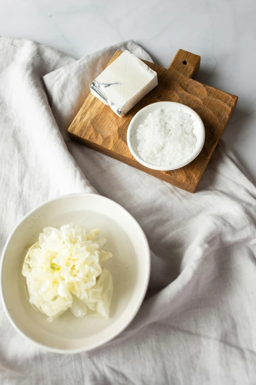 a white bowl sitting on top of a table next to a cutting board, a still life, carved soap, white petal, salt, lush