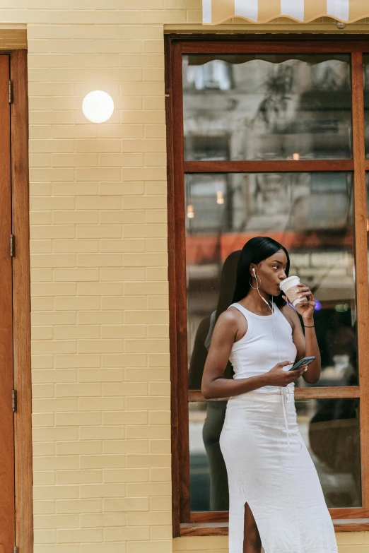 a woman in a white dress standing in front of a building, trending on unsplash, happening, coffee shop, dark skinned, drinking, light tan