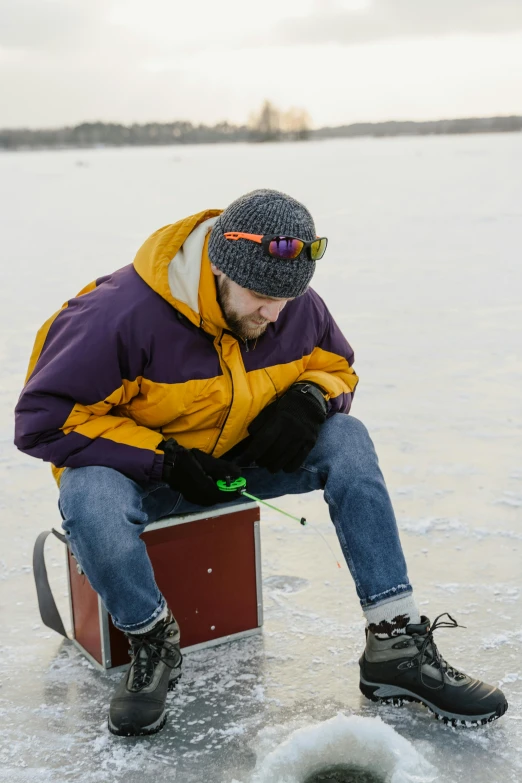 a man sitting on top of a box on a frozen lake, wearing a purple cap, fishing, warm features, yellow