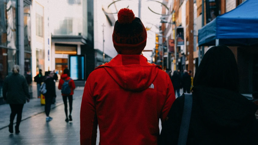 a person in a red jacket walking down a street, wearing a beanie, guide, unsplash photography, back of head