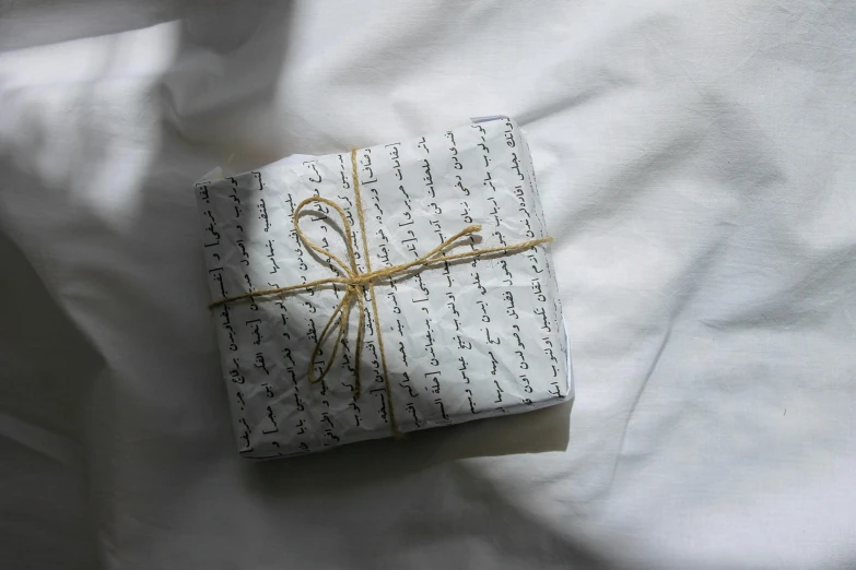 a wrapped gift sitting on top of a bed, an album cover, by Alice Mason, unsplash, hurufiyya, calligraphic poetry, white clothes, ignant, ancient writing