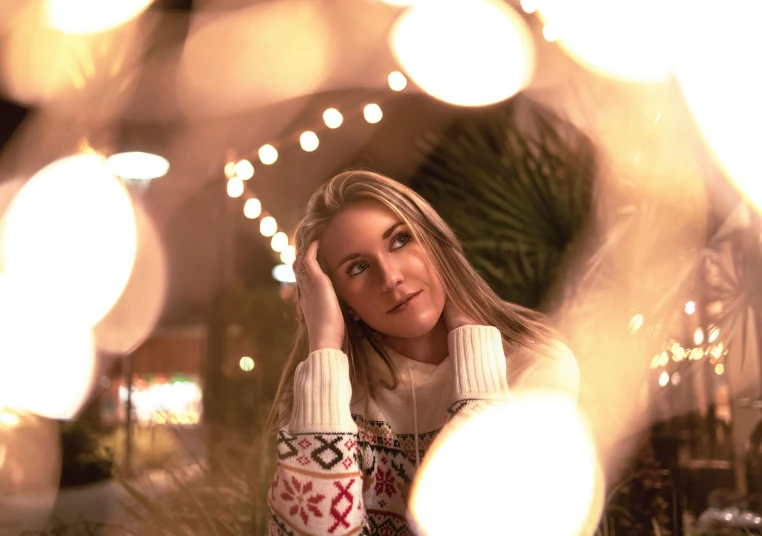 a woman sitting at a table with christmas lights in the background, a portrait, by Julia Pishtar, pexels contest winner, handsome girl, kaleidoscopic, a blond, gif