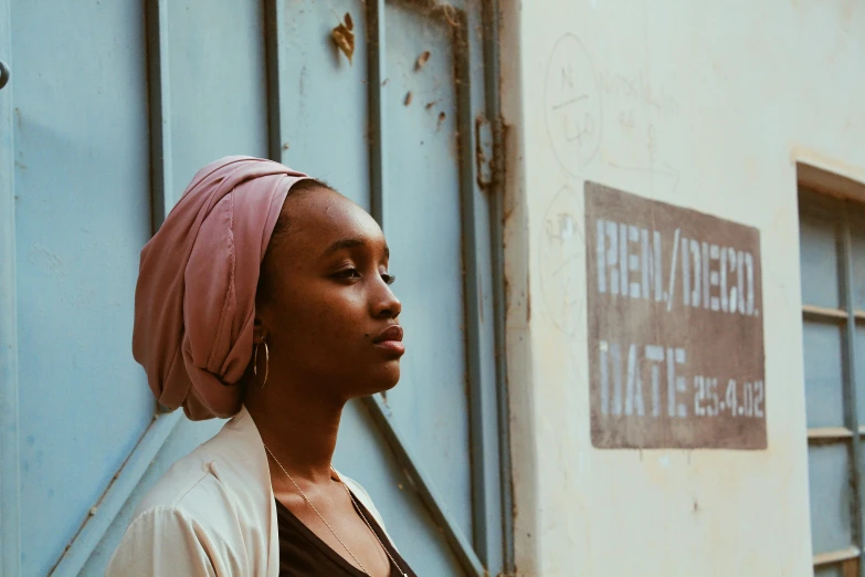 a woman standing in front of a blue door, a photo, by Matija Jama, pexels contest winner, afrofuturism, wearing a head scarf, wearing a pink head band, thoughtful ), ( brown skin )