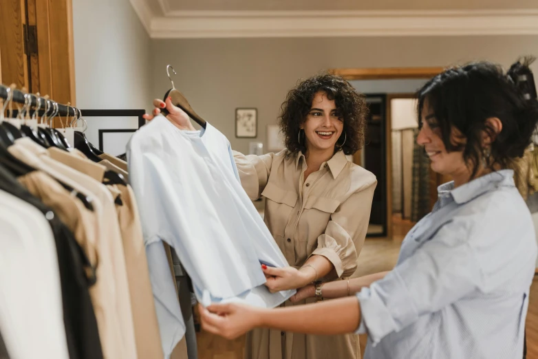 two women standing in front of a rack of clothes, pexels contest winner, wearing a linen shirt, avatar image, presenting wares, candid photo