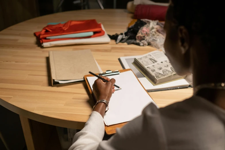 a woman sitting at a table writing on a piece of paper, a drawing, by Daniel Seghers, pexels contest winner, tailored clothing, wooden desks with books, kara walker, thumbnail