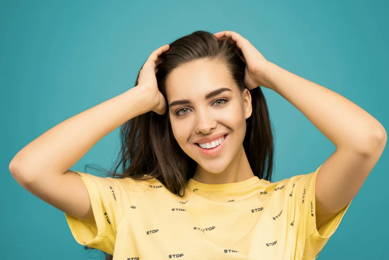 a woman holding her hair in front of her head, trending on pexels, hurufiyya, wearing a modern yellow tshirt, attractive brown hair woman, perfect teeth, photoshoot for skincare brand