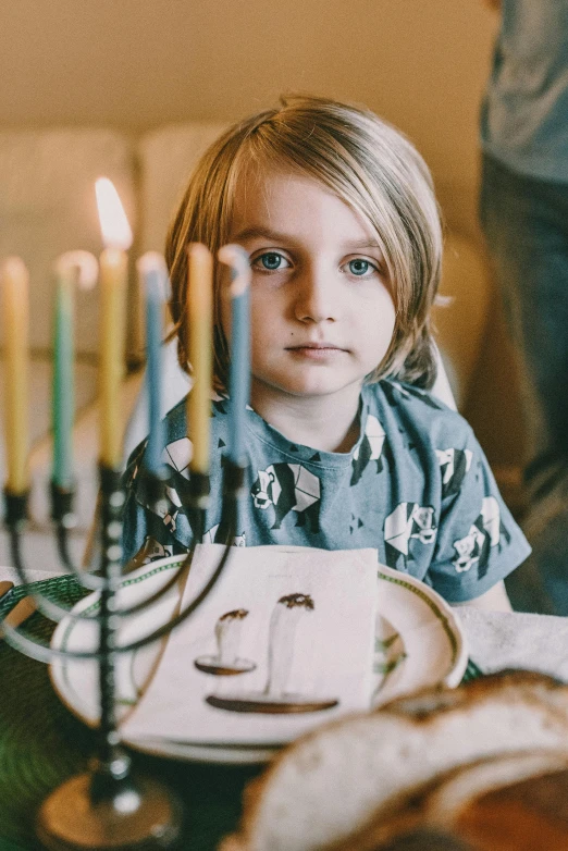 a little girl sitting at a table with a menorah, a colorized photo, pexels, blonde boy with yellow eyes, wearing festive clothing, portrait featured on unsplash, at a dinner table