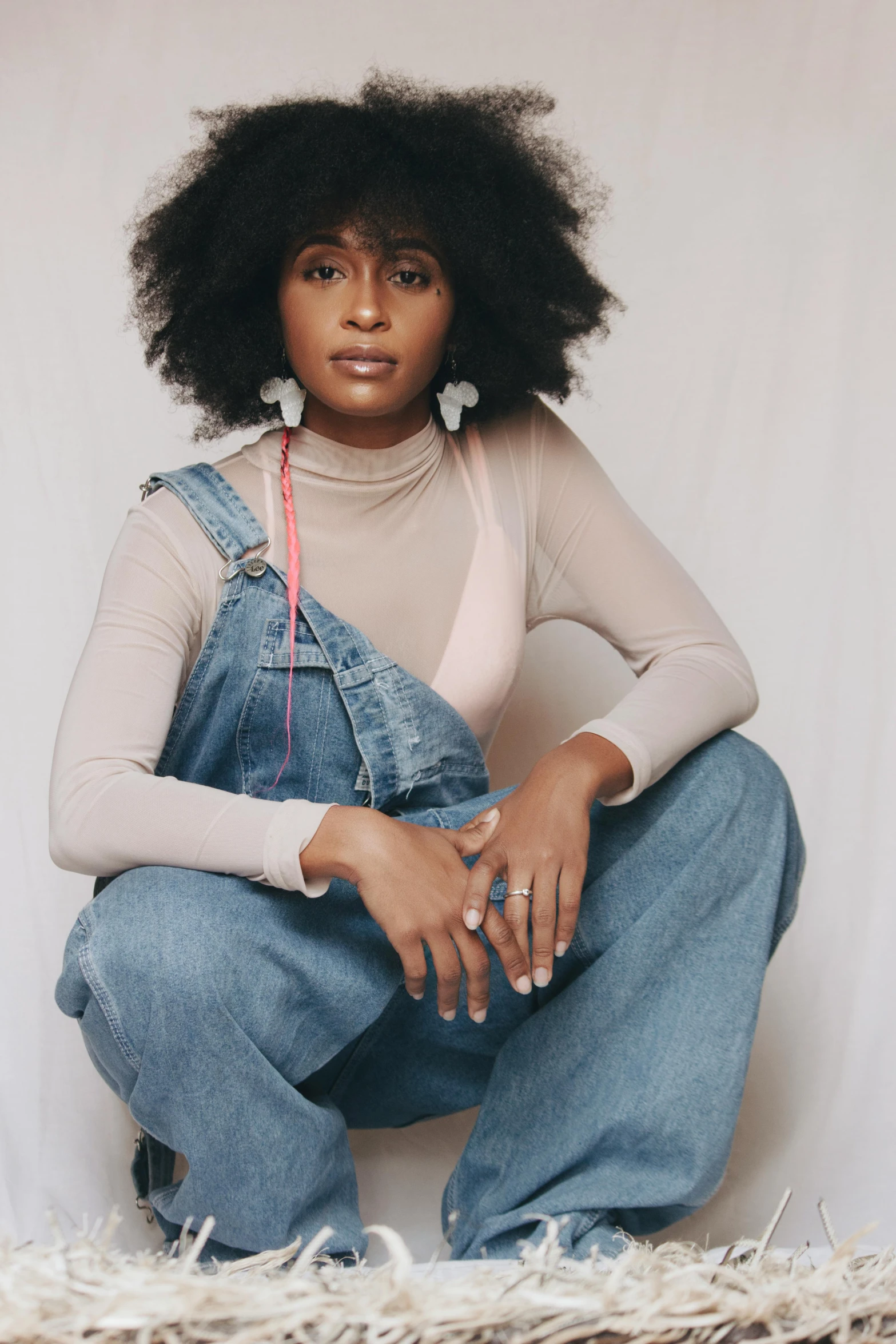 a woman sitting on the floor wearing overalls, by Lily Delissa Joseph, trending on pexels, afrofuturism, long afro hair, blue turtleneck, wearing several pendants, full body and head shot