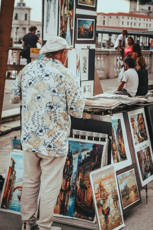 a man standing next to a table with pictures on it, a fine art painting, pexels contest winner, street market, facing away, 4k photograph of painting, patterned clothing