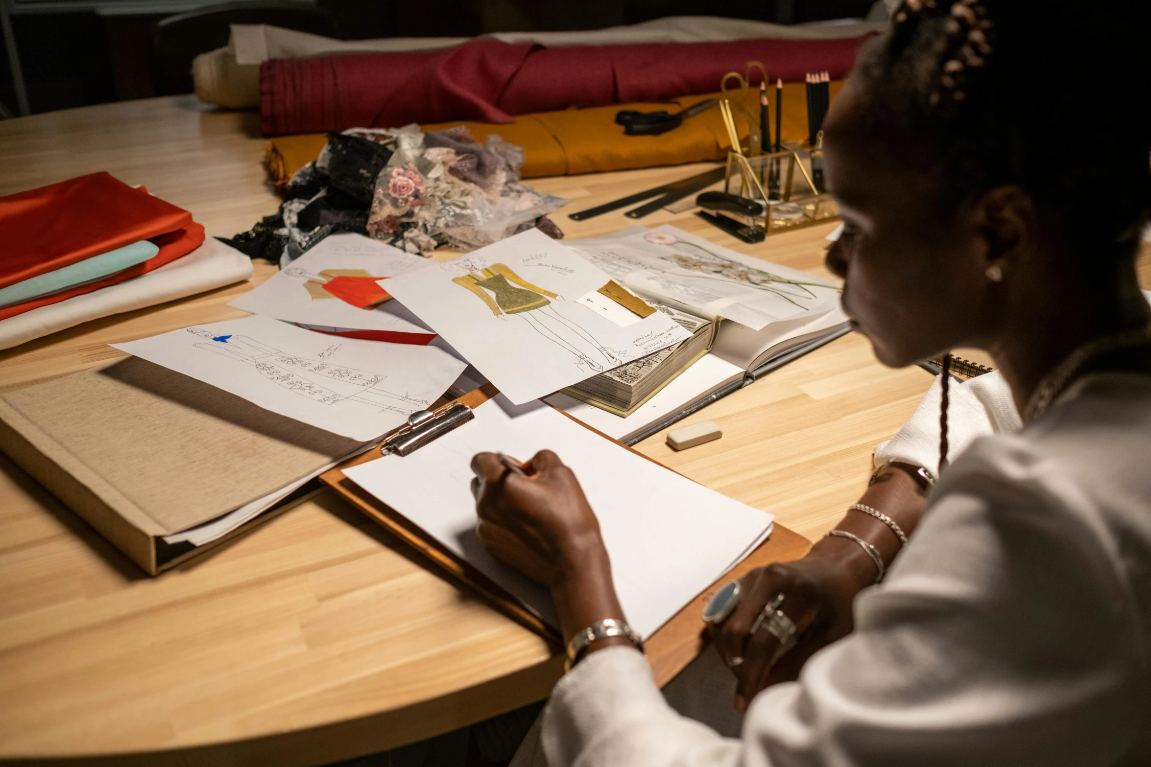 a woman sitting at a table writing on a piece of paper, a drawing, inspired by Afewerk Tekle, pexels contest winner, fashion designer, stitching, brown, in the evening