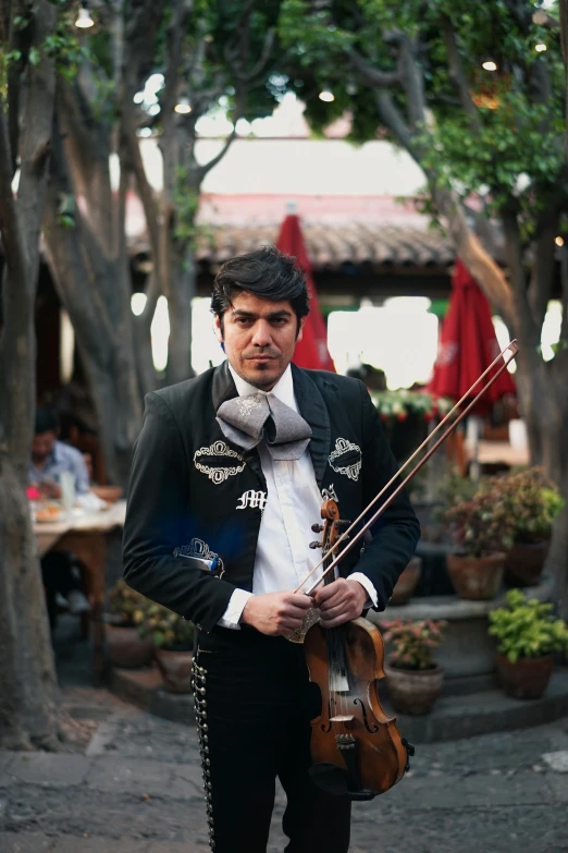 a man standing in a courtyard holding a violin, an album cover, by Alejandro Obregón, standing in a restaurant, wearing an ornate outfit, trending photo, mexican standoff