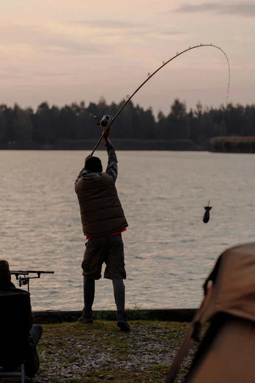 a man standing next to a body of water holding a fishing pole, in the evening, big crowd, outdoors, hanging