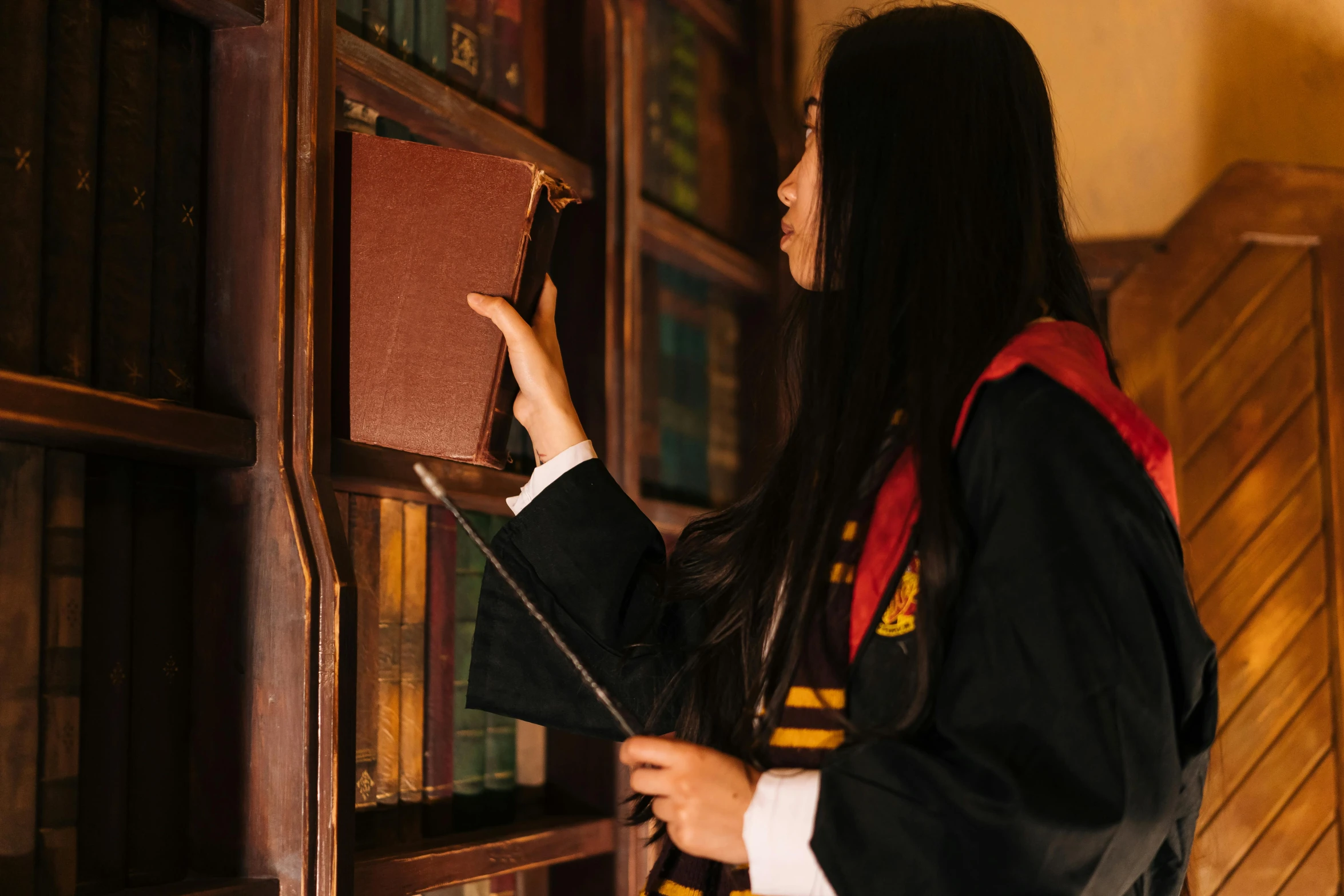 a woman standing in front of a bookshelf holding a book, inspired by Hermione Hammond, pexels contest winner, academic art, hogwarts gryffindor common room, wearing an academic gown, seifuku, maintenance photo