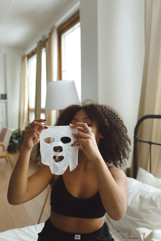 a woman sitting on a bed holding a paper mask in front of her face, glowing skin face, with afro, silicone skin, skimask