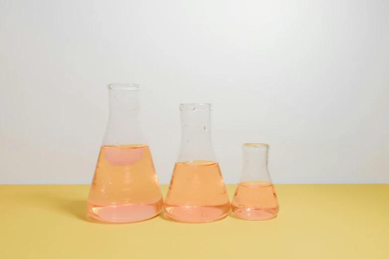three flasks sitting next to each other on a yellow surface, unsplash, hyperrealism, featuring pink brains, scientific glassware, in triangular formation, light orange mist