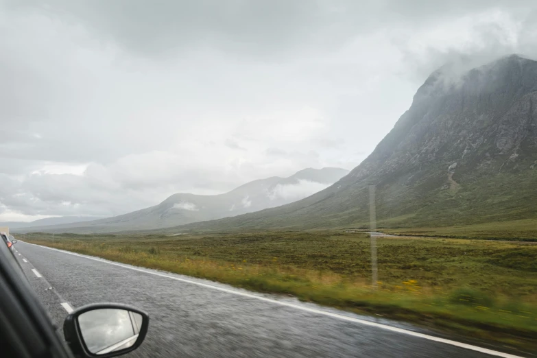 a car driving down a road next to a mountain, by Roar Kjernstad, hurufiyya, grey skies rain, biggish nose, thumbnail, panoramic shot