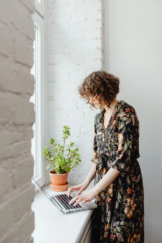 a woman standing in front of a window using a laptop, by Nicolette Macnamara, potted plant, curly haired, floral patterned skin, professionally