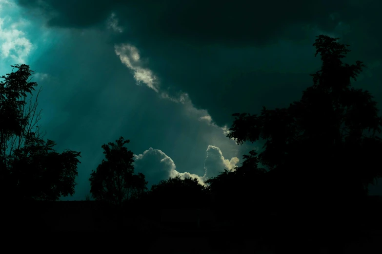 a dark sky with clouds and trees in the foreground, inspired by Elsa Bleda, pexels contest winner, magical realism, rays of sunlight, dramatic clouds cyan atmosphere, silhouetted, god ray