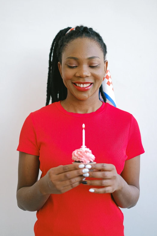 a woman in a red shirt holding a cupcake, white candles, dark skinned, wearing a party hat, promo image