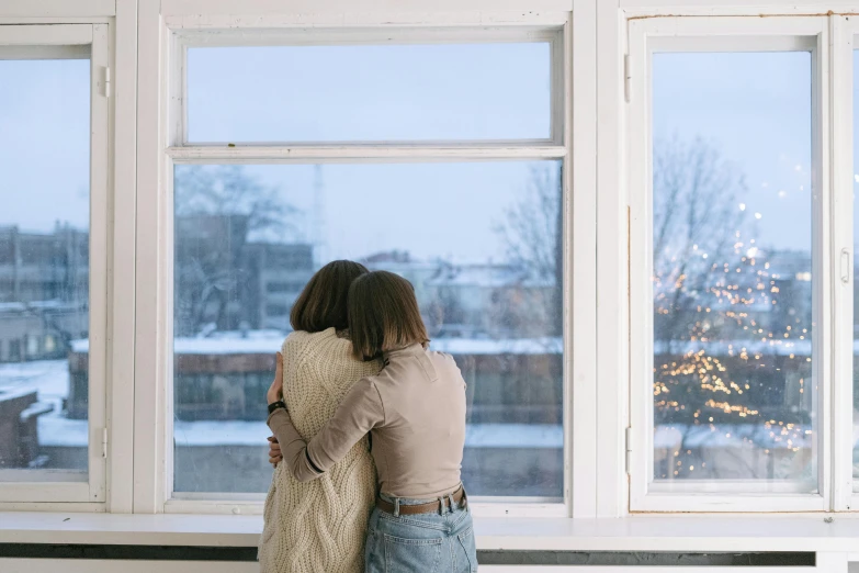 two women hugging each other in front of a window, trending on pexels, winter season, background image, lonely, lgbtq