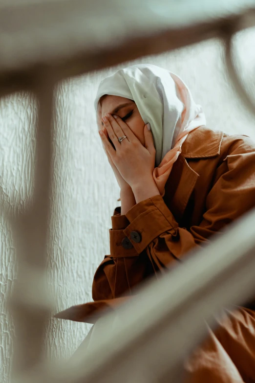 a woman covers her face with her hands, a picture, trending on pexels, hurufiyya, white hijab, tired expression, stood in a cell, brown clothes