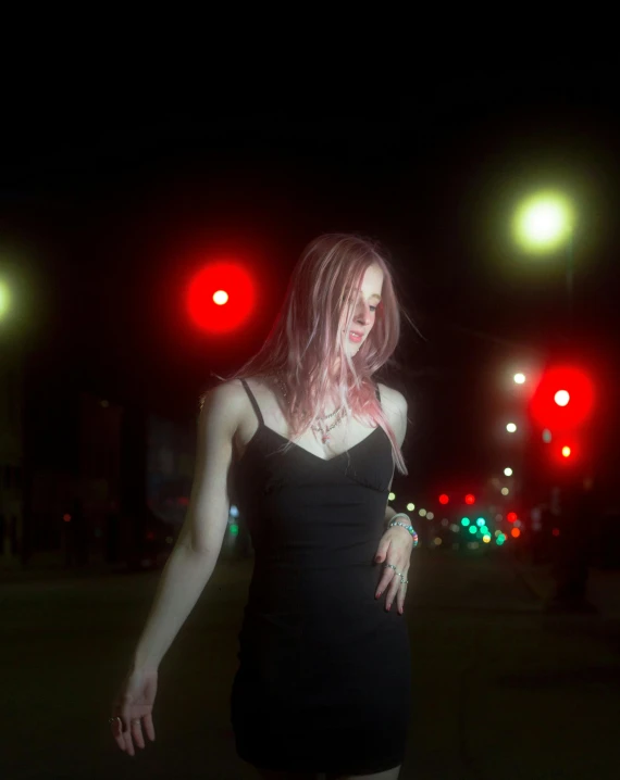 a woman standing in the middle of a street at night, an album cover, unsplash, neo-figurative, albino white pale skin, red lights, non binary model, grungy woman with rainbow hair