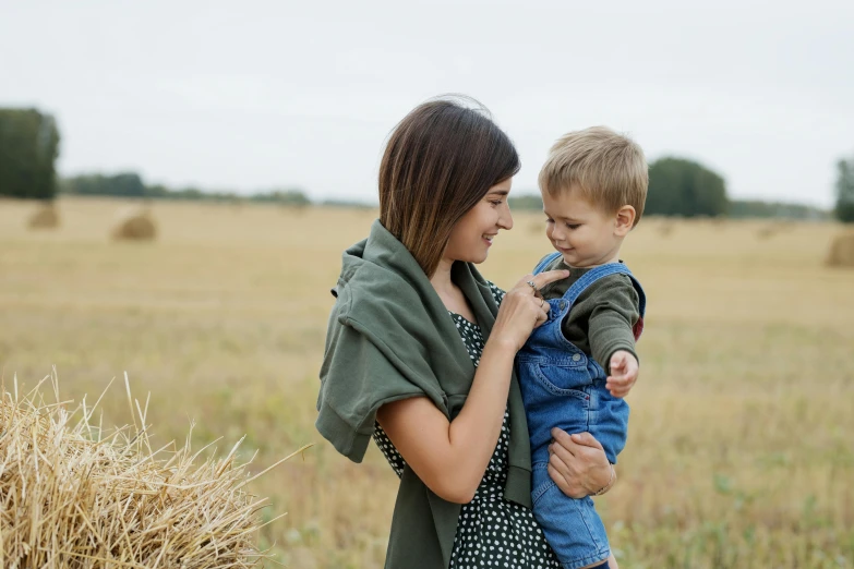 a woman holding a small child in a field, pexels contest winner, wearing hay coat, overalls, stubble, high quality product image”