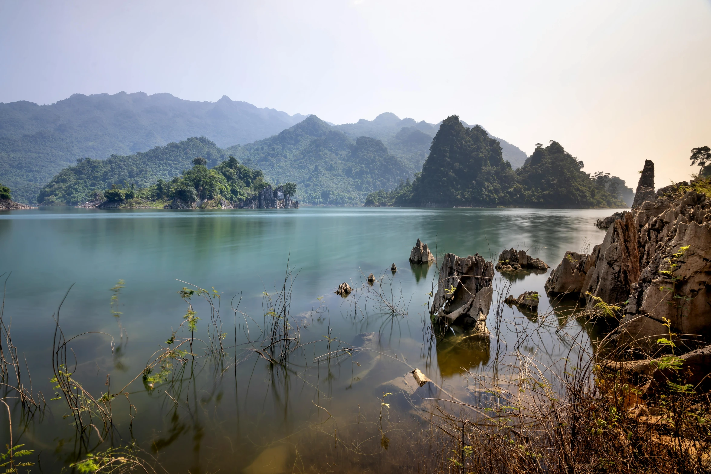 a large body of water surrounded by trees, by Tom Wänerstrand, unsplash contest winner, sumatraism, vietnam, mountains in background, floating magical rocks, in the middle of a lake