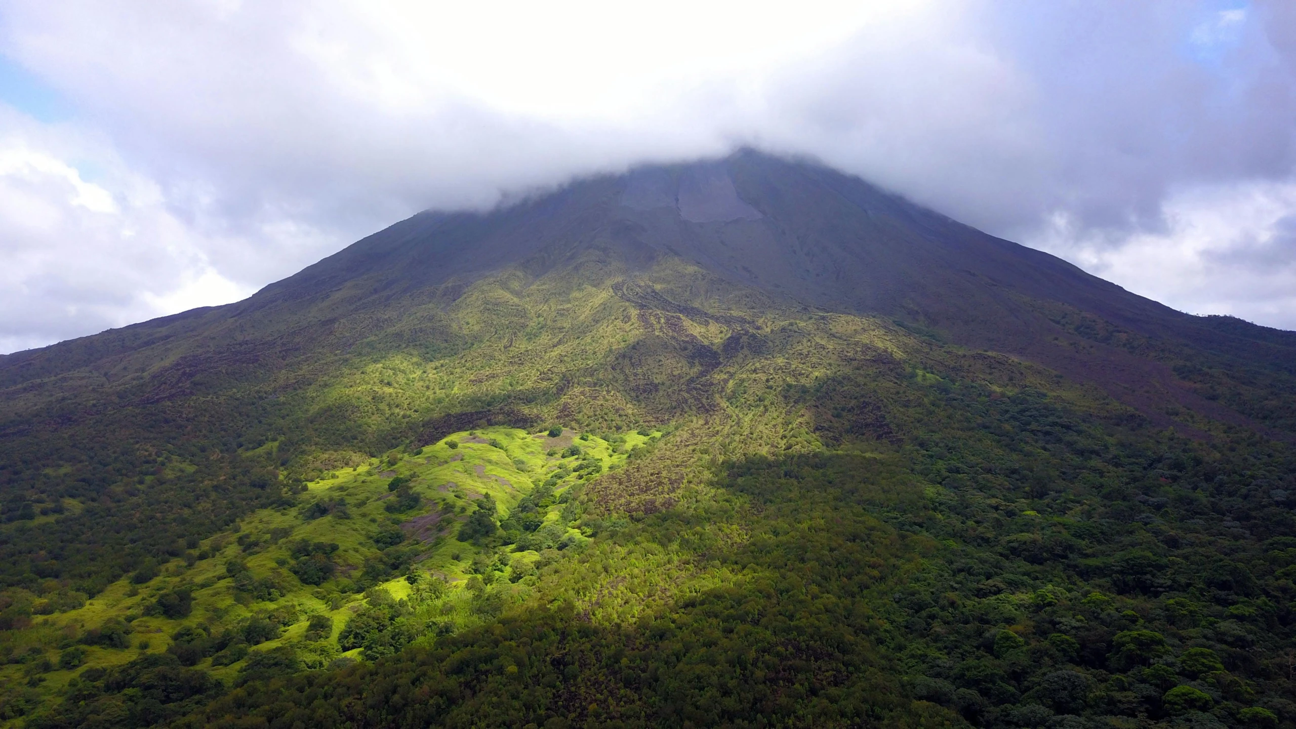 an aerial view of a mountain on a cloudy day, by Carey Morris, hurufiyya, in a volcano, verdant and lush and overgrown, conde nast traveler photo, drone footage