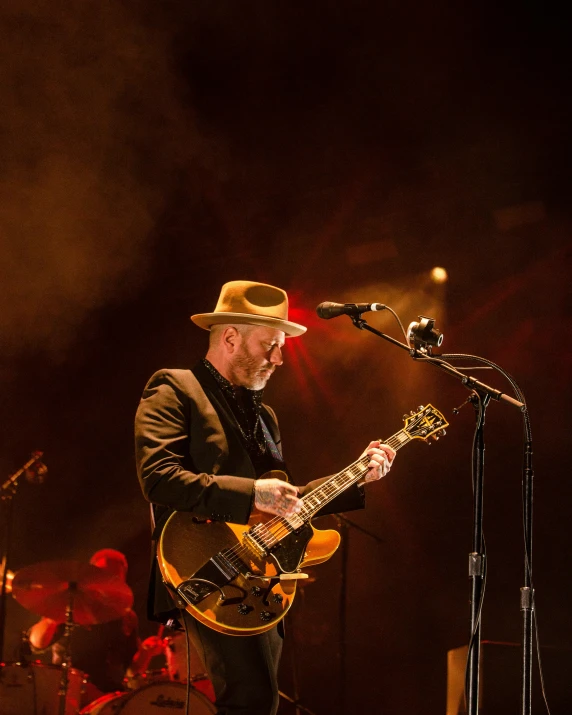 a man that is playing a guitar on a stage, tipping his fedora, colin hay, profile image, paul giamatti