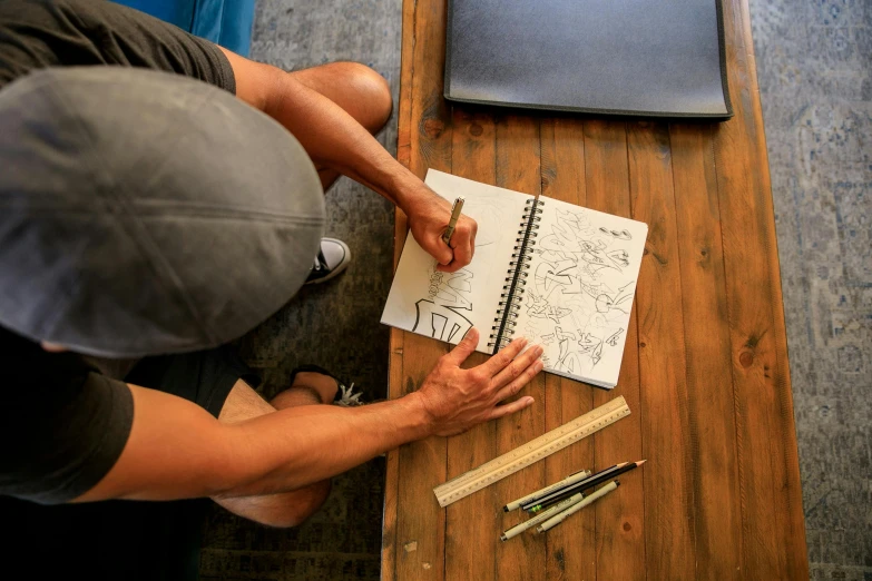 a man sitting at a table drawing on a piece of paper, a drawing, by Meredith Dillman, pexels contest winner, holding notebook, aaron horkey, thumbnail, a high angle shot