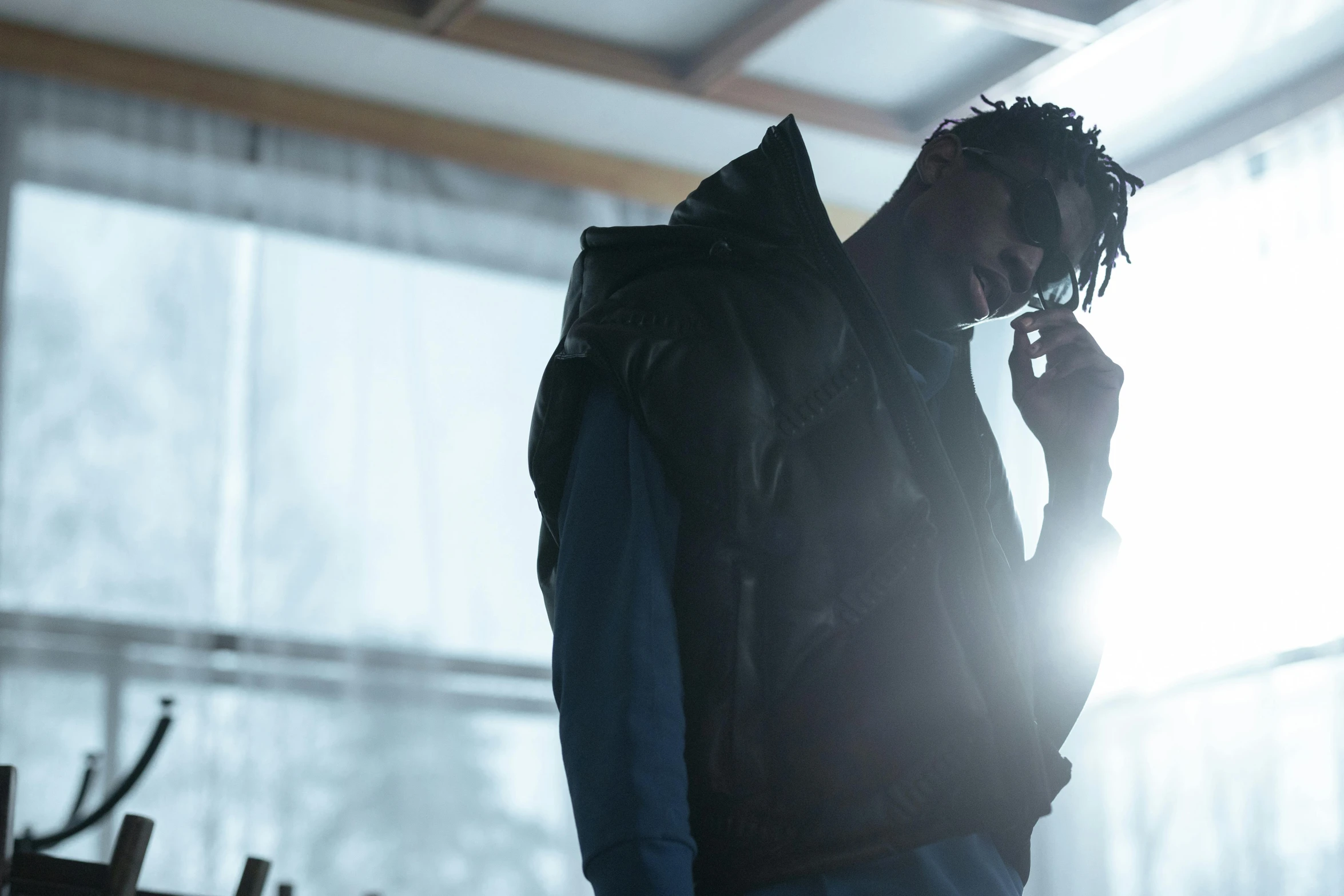 a man standing in front of a window talking on a cell phone, inspired by Liam Wong, unsplash, afrofuturism, he wears an eyepatch, backlit shot girl in parka, 2 1 savage, standing in a dimly lit room