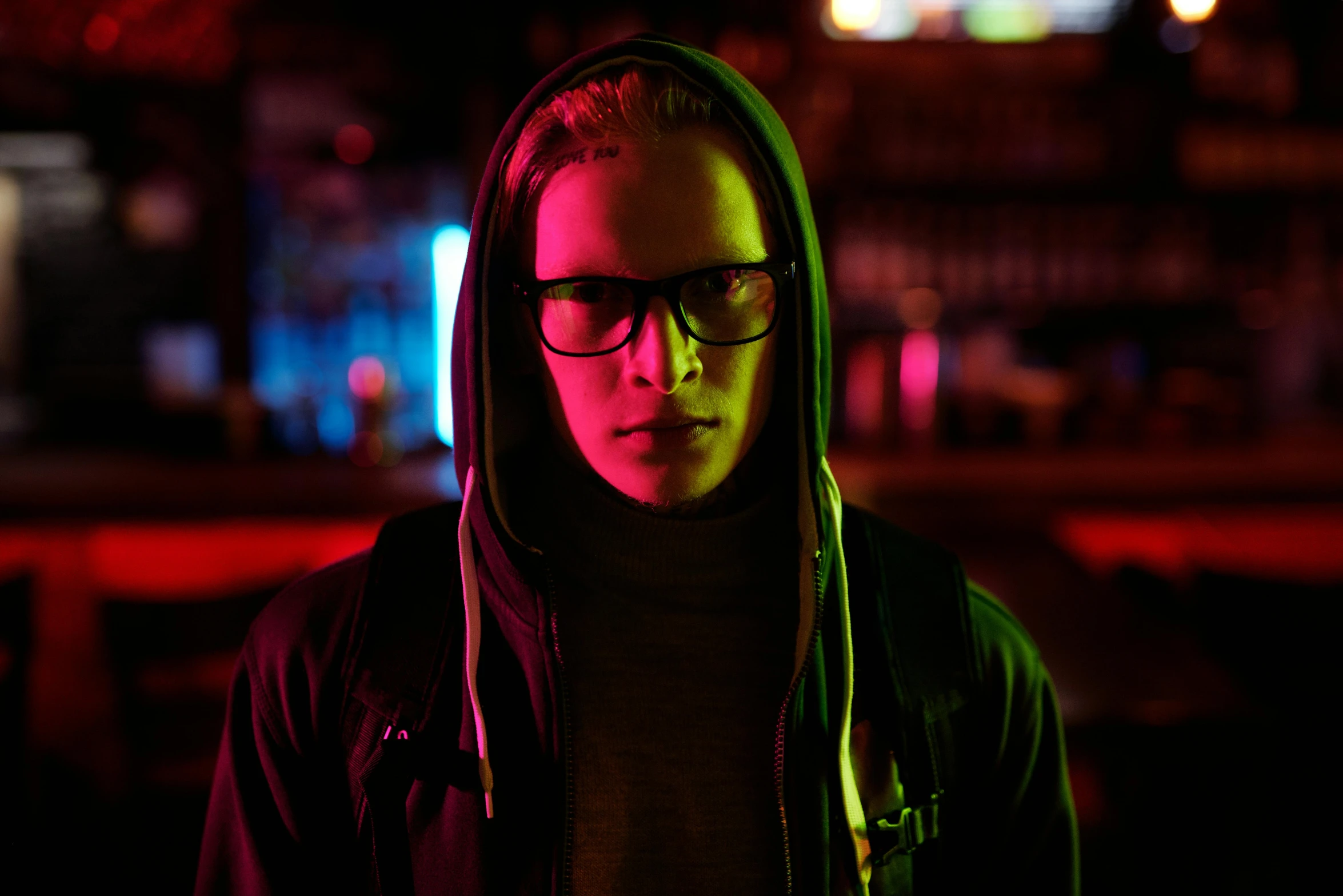 a man with glasses and a hoodie standing in front of a bar, cyberpunk art, pexels, bisexual lighting, filmic lut, teenage boy, quicksilver