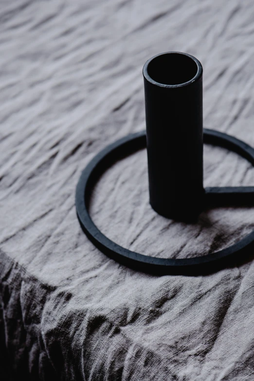 a black candle holder sitting on top of a bed, an abstract sculpture, by Nina Hamnett, unsplash, minimalism, circle face, simple primitive tube shape, cast iron material, detailed shot