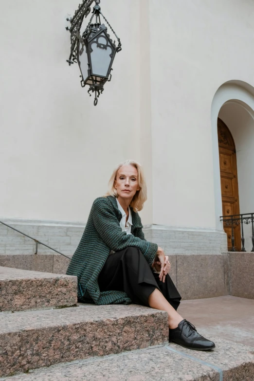 a woman sitting on the steps of a building, by Grytė Pintukaitė, trending on unsplash, wearing a cardigan, russian academic, casual green clothing, blonde swedish woman