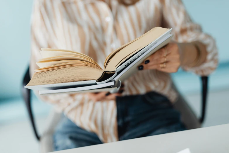 a woman sitting in a chair holding an open book, pexels contest winner, sleek spines, on a white table, reading engineering book, curated collections