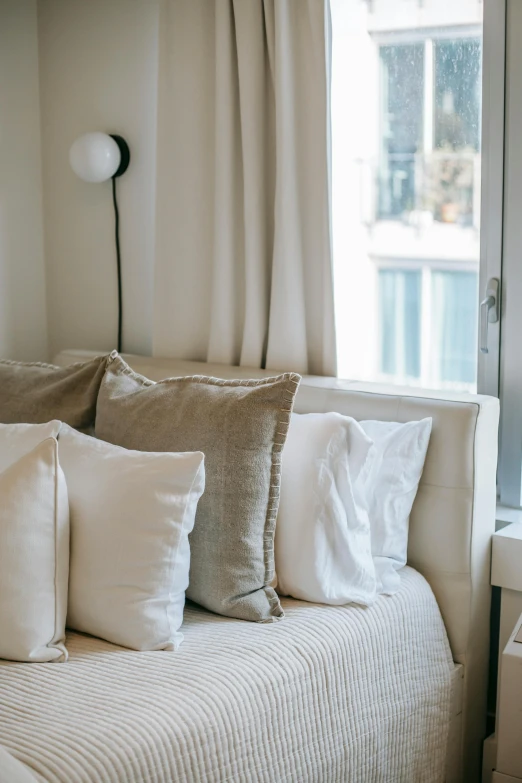 a bed sitting in a bedroom next to a window, by William Home Lizars, trending on unsplash, light and space, light beige pillows, white l shaped couch, silver，ivory, city apartment cozy calm