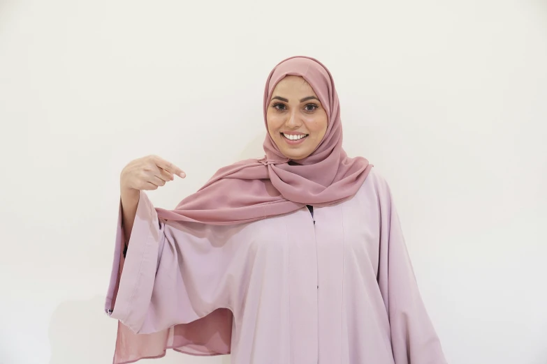 a woman in a hijab poses for a picture, pexels contest winner, hurufiyya, light pink, royal cape, plain background, arms extended