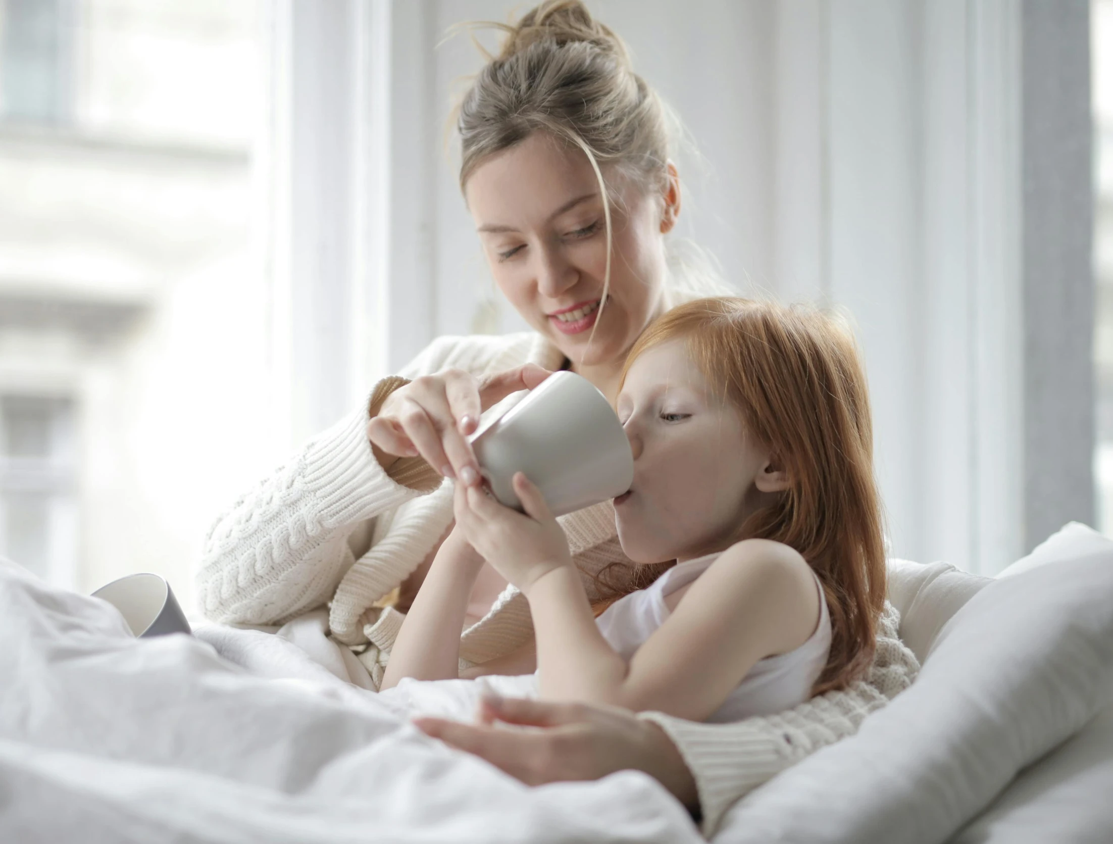 a woman sitting on top of a bed next to a little girl, is ((drinking a cup of tea)), grey, gen z, manuka