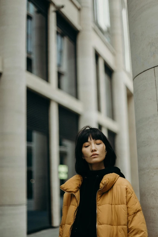 a woman standing in front of a tall building, inspired by Wang E, pexels contest winner, muted brown yellow and blacks, gemma chan girl portrait, she wears a jacket, ethnicity : japanese